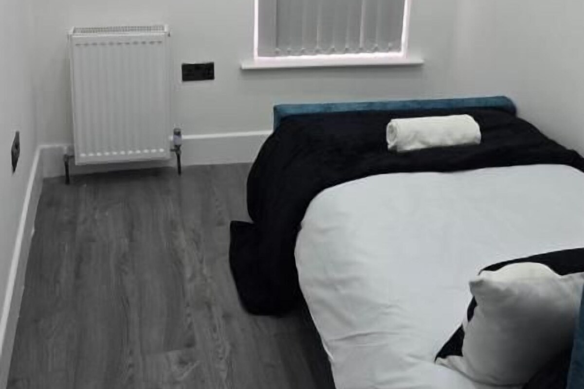 A bedroom with a bed, a radiator, a window with blinds, and a cityscape painting on the wall.