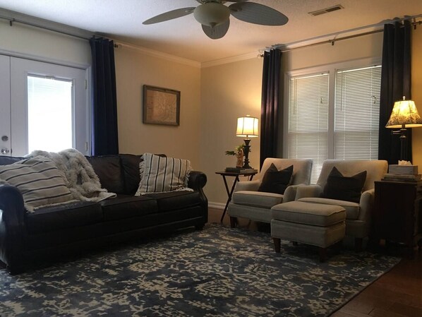 Een woonkamer met een donkere bank, twee fauteuils, een vloerkleed met een patroon, een plafondventilator en een lamp.