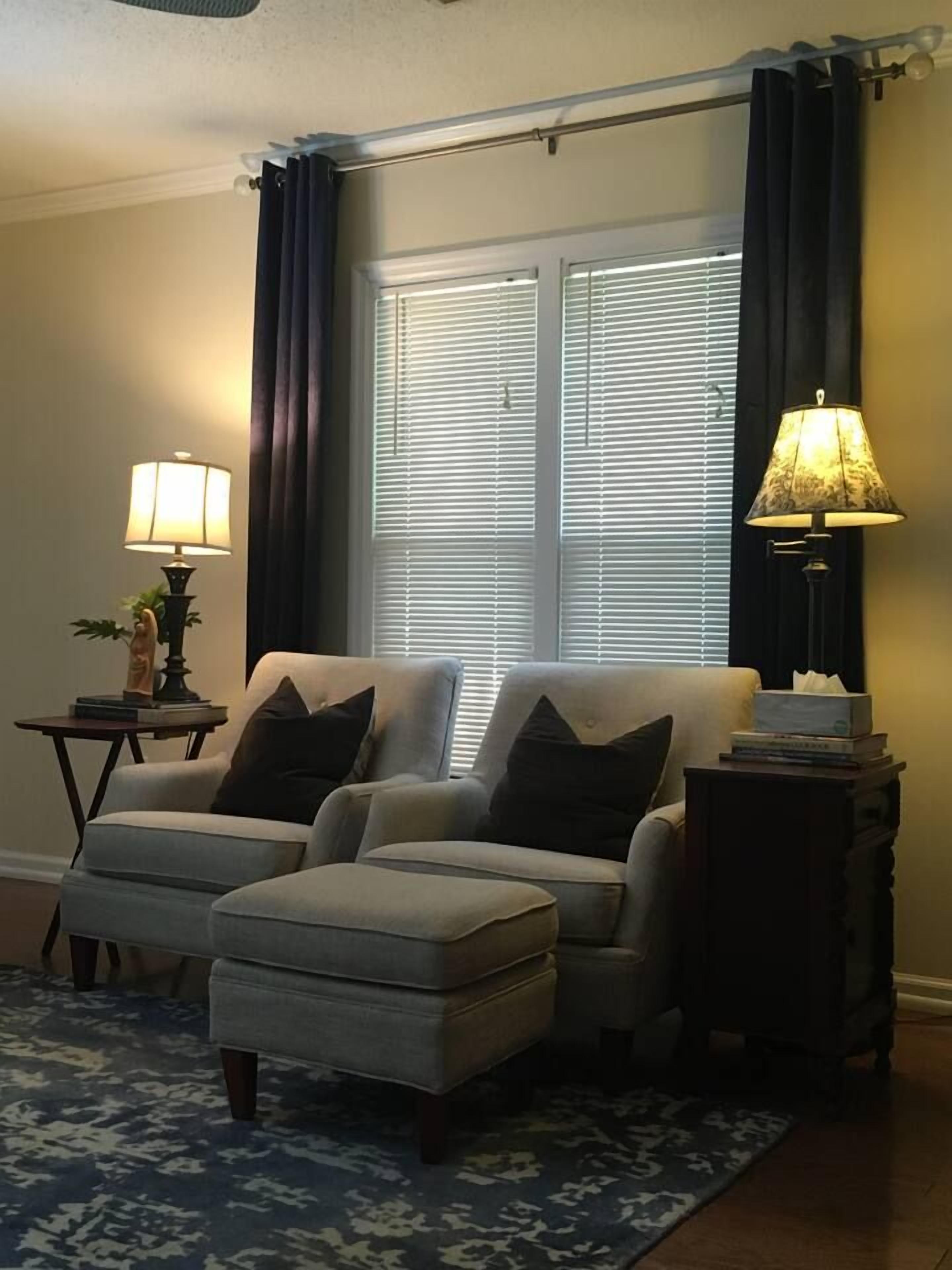 A living room with two armchairs, a coffee table, and a rug. There are two lamps on side tables and a window with blinds.