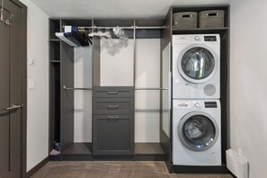 A laundry room with a built-in washer and dryer, a shelving unit with hanging clothes, and a door with a towel rack. - Downtown | On Bus Route | Garage Gear Storage | Pet Friendly (Steamboat Springs)