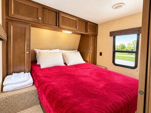 A bedroom with a red bedspread, white pillows, and a view of a green landscape through a window. - Cozy RV Getaway in Sturgis Private Bedroom Bunk beds Full Kitchen (Sturgis)