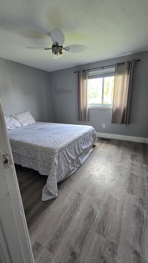 Ein Schlafzimmer mit einem Bett, einem Deckenventilator, einem Fenster mit Vorhängen und Holzboden.