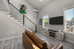 A modern living room with a staircase, a mounted television, and a large window. - Carriage House: No Cleaning/Pet Fees Soaking Tub Pet Friendly (Galveston)