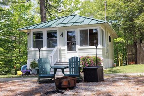 A small, single-story cabin with a green roof, numbered 4, featuring a porch with two chairs and a table, surrounded by a gravel driveway and trees. - Lavish Little Lodge (Conway)
