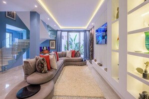 A modern living room with a curved sofa, a glass staircase, and a view of greenery outside. - Modern 4-bedroom villa with a pool and hot tub - Golf Community (Marrakech)