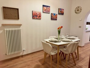 Ein Essbereich mit einem weißen Tisch, der für vier Personen gedeckt ist, umgeben von weißen Stühlen. An der Wand über dem Tisch hängen vier gerahmte Gemälde.