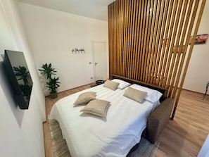 Ein modernes Schlafzimmer mit Bett, Nachttischen, einem Spiegel, einer Topfpflanze und einem Raumteiler aus Holzlatten.