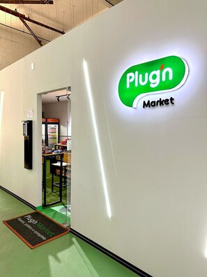 A corridor with a wall-mounted sign reading 'Plug Market' and a door leading to a room with a vending machine and a table. - Maia Supreme Apartment 012207 5 min Shopping Maia (Guarulhos)