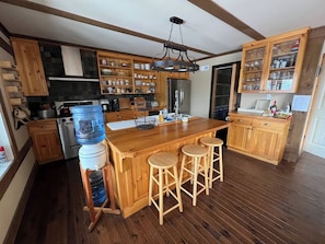 A kitchen with wooden cabinets, a central island with stools, a water dispenser, and a hanging light fixture.