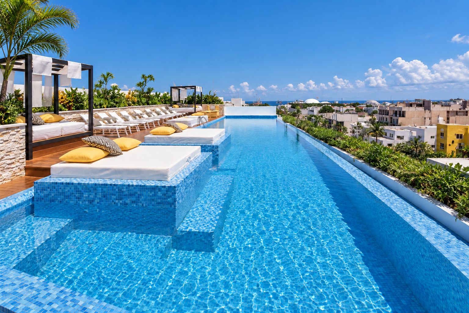 A long swimming pool with lounge chairs and sunbeds along the side, surrounded by greenery and buildings.