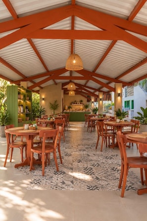 Un comedor interior con mesas y sillas de madera, suelo estampado y techo alto con vigas de madera.
