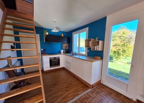 A kitchen with a wooden staircase, white cabinets, a built-in oven, and a large window with a view of greenery. - Getaway in Northern Brittany: House with a garden, 1 km from the sea and with access to the GR34 trail (Plougasnou)