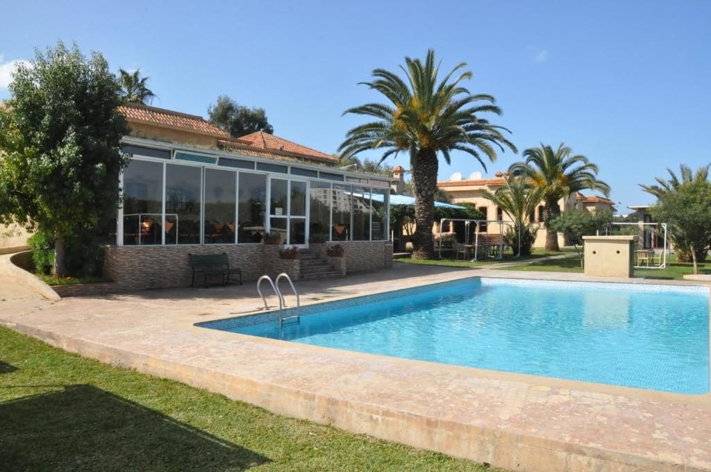 A swimming pool with a glass-enclosed patio area, surrounded by palm trees and a building with a tiled roof.