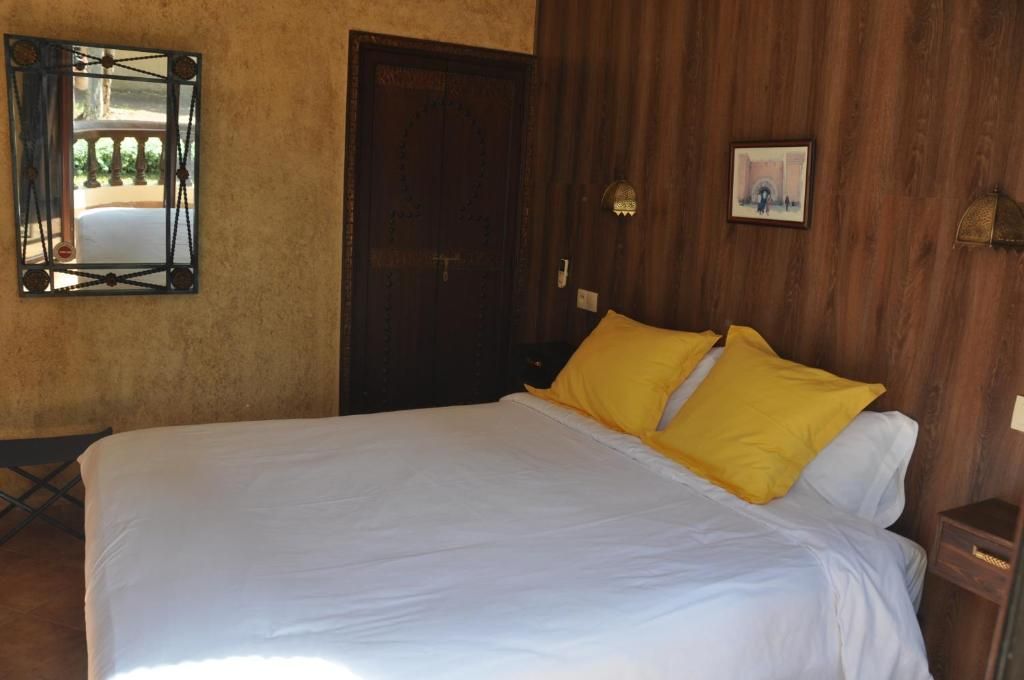 A bed with white bedding and two yellow pillows, a wooden door, a small bedside table, and a wall-mounted light fixture.