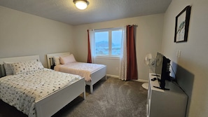 Ein Schlafzimmer mit zwei Betten, einem fensterbelaubten Fenster, einem Schreibtisch mit Computer und einem am Wand montierten Fernseher.
