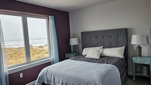 Ein Schlafzimmer mit großem Fenster, einem Bett mit grauem Kopfteil, zwei Nachttischen mit Lampen und Blick auf den Strand.