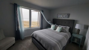 Ein Schlafzimmer mit einem Bett, einem Nachttisch, einer Lampe, einem Fenster mit Blick auf den Strand und einem Bild an der Wand.