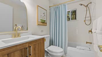 Een badkamer met een witte wastafel, gouden kraanwerk, een houten kastje, een wit toilet, een bad met douchekop en een blauw douchegordijn.
