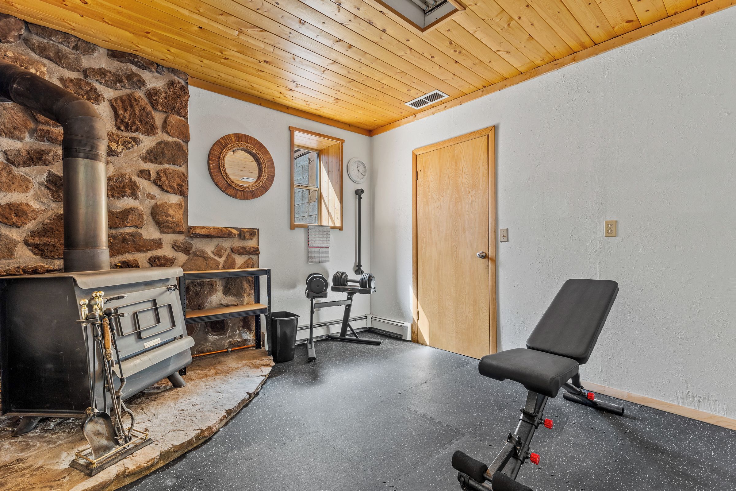 A room with a stone wall, a wooden ceiling, a black exercise chair, a door, and a mirror.