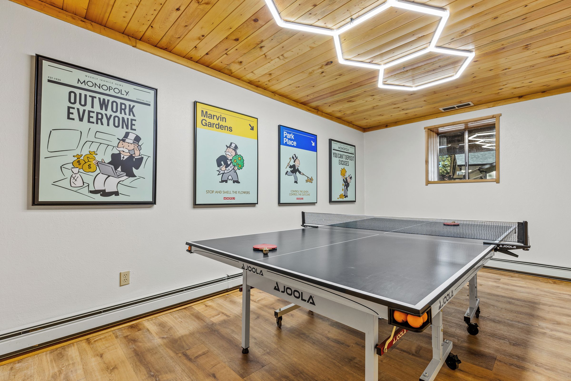 Una mesa de ping pong en una habitación con suelo de madera, un techo con luces hexagonales y cuatro carteles con temática de Monopoly en la pared.