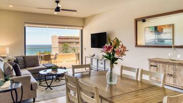 A spacious living area with a balcony overlooking the sea, a dining table, and a TV mounted on the wall.
