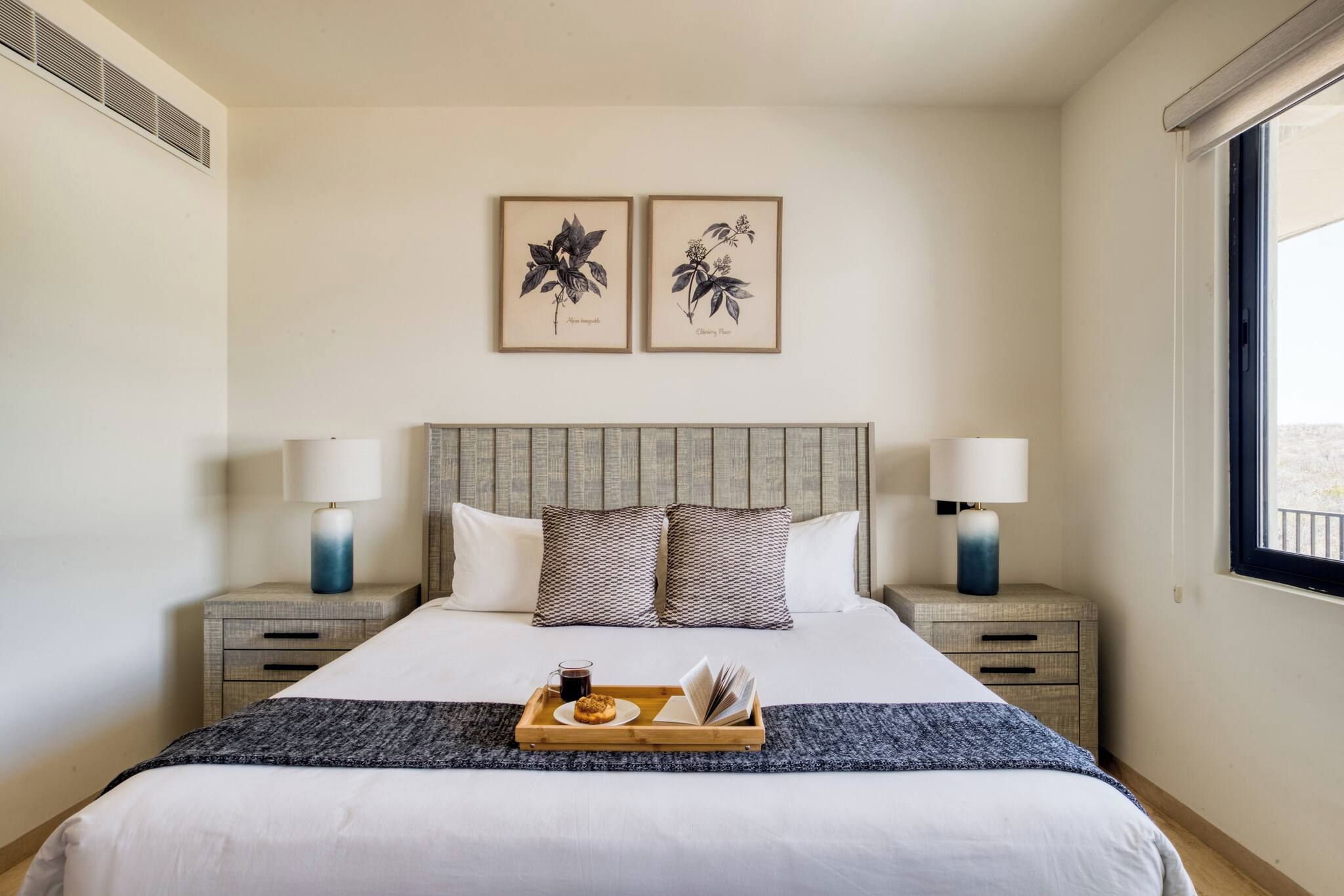 A neatly arranged hotel room with a large bed, two bedside tables with lamps, a tray with a muffin and a cup, and two framed botanical prints on the wall.