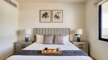 A neatly arranged hotel room with a large bed, two bedside tables with lamps, a tray with a muffin and a cup, and two framed botanical prints on the wall.