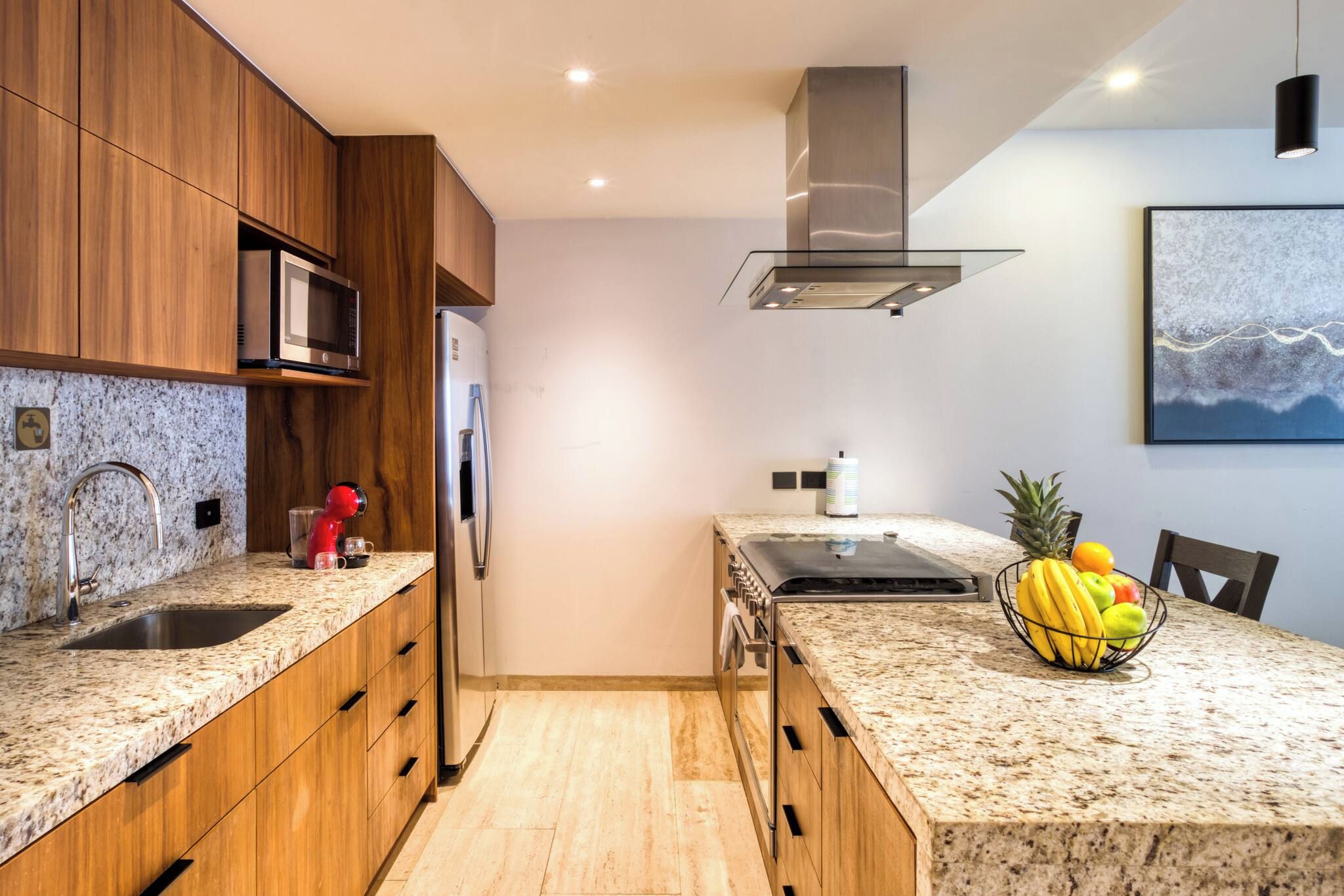 Una cocina moderna con gabinetes de madera, electrodomésticos de acero inoxidable, encimeras de granito y un frutero en la isla.