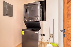 Una unidad compacta de lavadora y secadora instalada en una habitación pequeña con una puerta de madera y una escoba apoyada contra la pared.