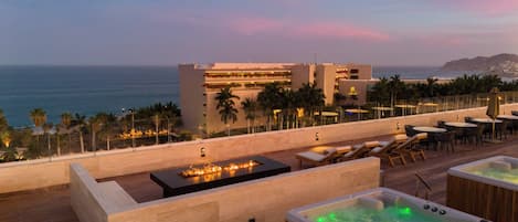 A rooftop terrace with a pool, lounge chairs, and a view of the ocean and a hotel building.