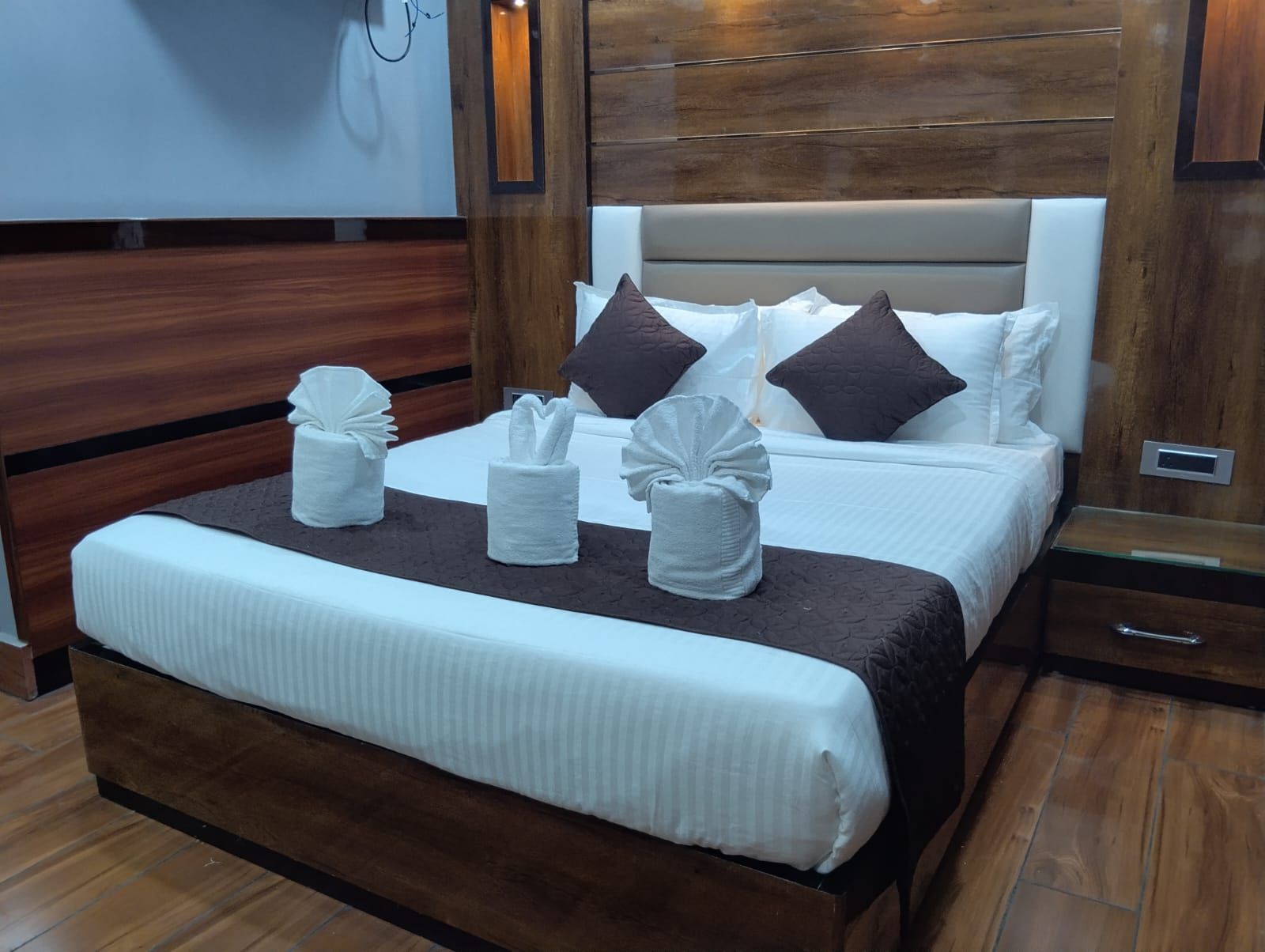Un lit bien fait, avec du linge de lit blanc et des serviettes décoratives, une tête de lit en bois et une table de chevet sur laquelle se trouve une horloge numérique.
