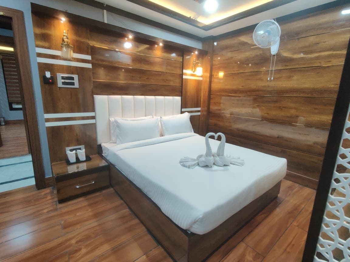 Une chambre d’hôtel moderne avec une tête de lit en bois, un lit avec du linge de maison blanc, une table de chevet avec une lampe et un ventilateur fixé au mur.