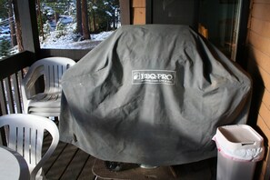 A covered grill with a logo, a white plastic chair, and a trash bin on a wooden deck. - Mammoth Love Nest – Mammoth Lakes (Mammoth Lakes)