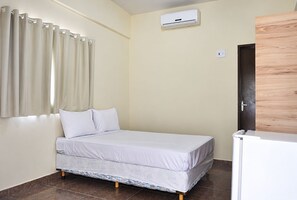 A single bed with a white mattress and pillows, a wall-mounted air conditioner, and a door with a wooden panel.