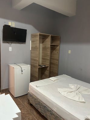 A compact room with a bed, a wooden wardrobe, a small white appliance, and a mounted television.