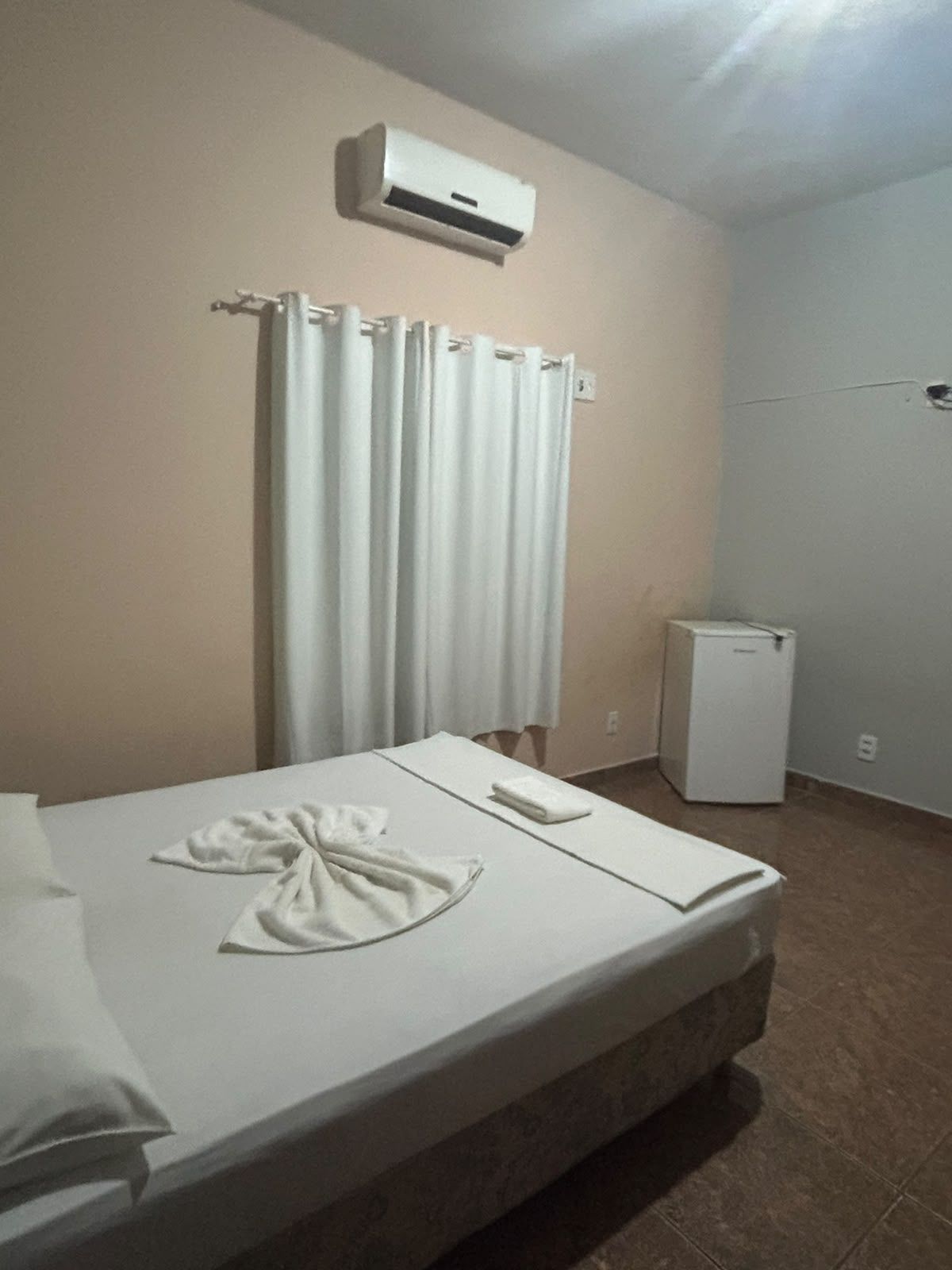 Una habitación de hotel con una cama, un refrigerador pequeño, un equipo de aire acondicionado y una cortina.