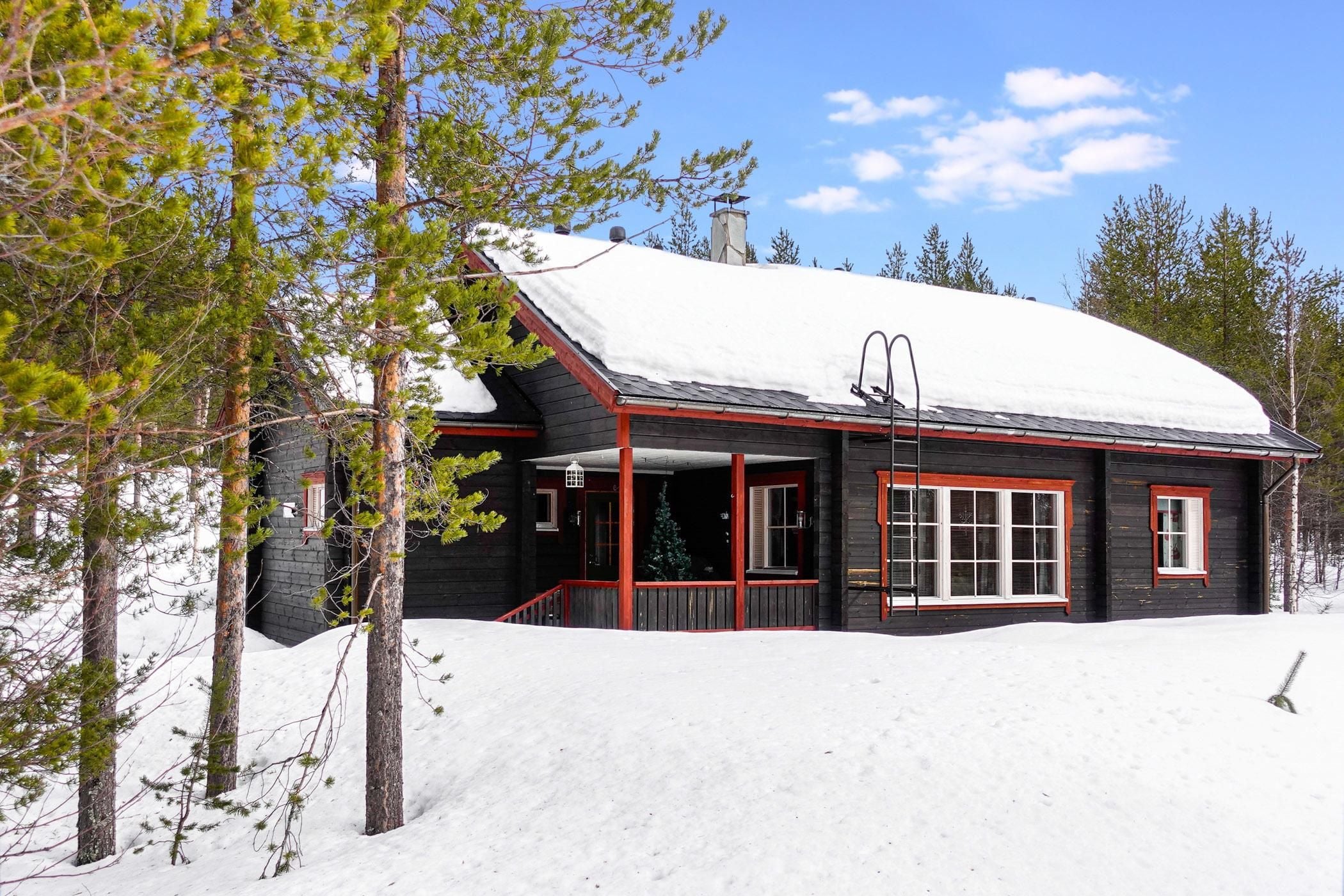 Una cabaña con techo cubierto de nieve, paredes de madera y un porche con detalles rojos.