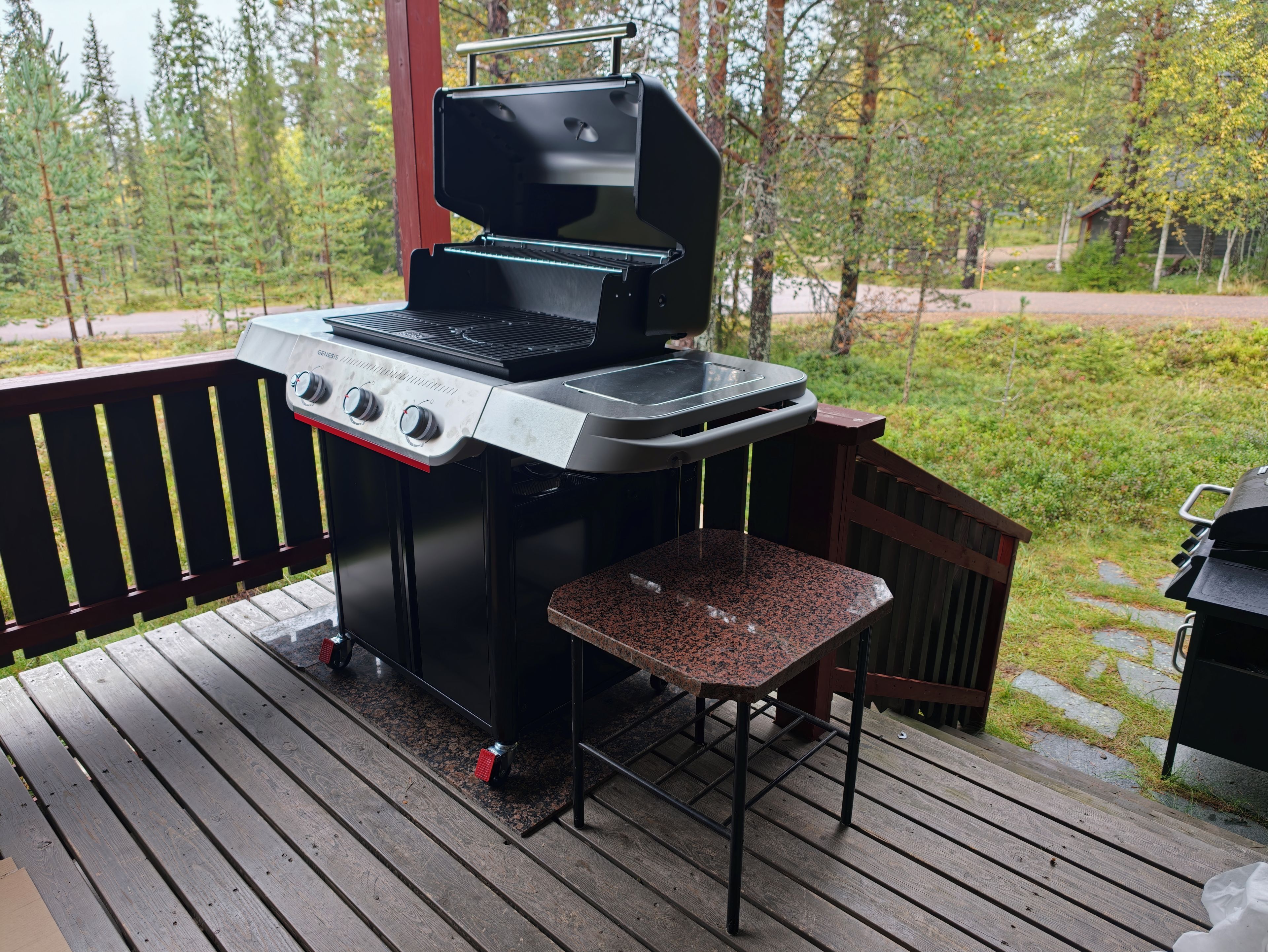 Una parrilla de barbacoa en una terraza exterior con barandilla de madera y vista a zona boscosa.