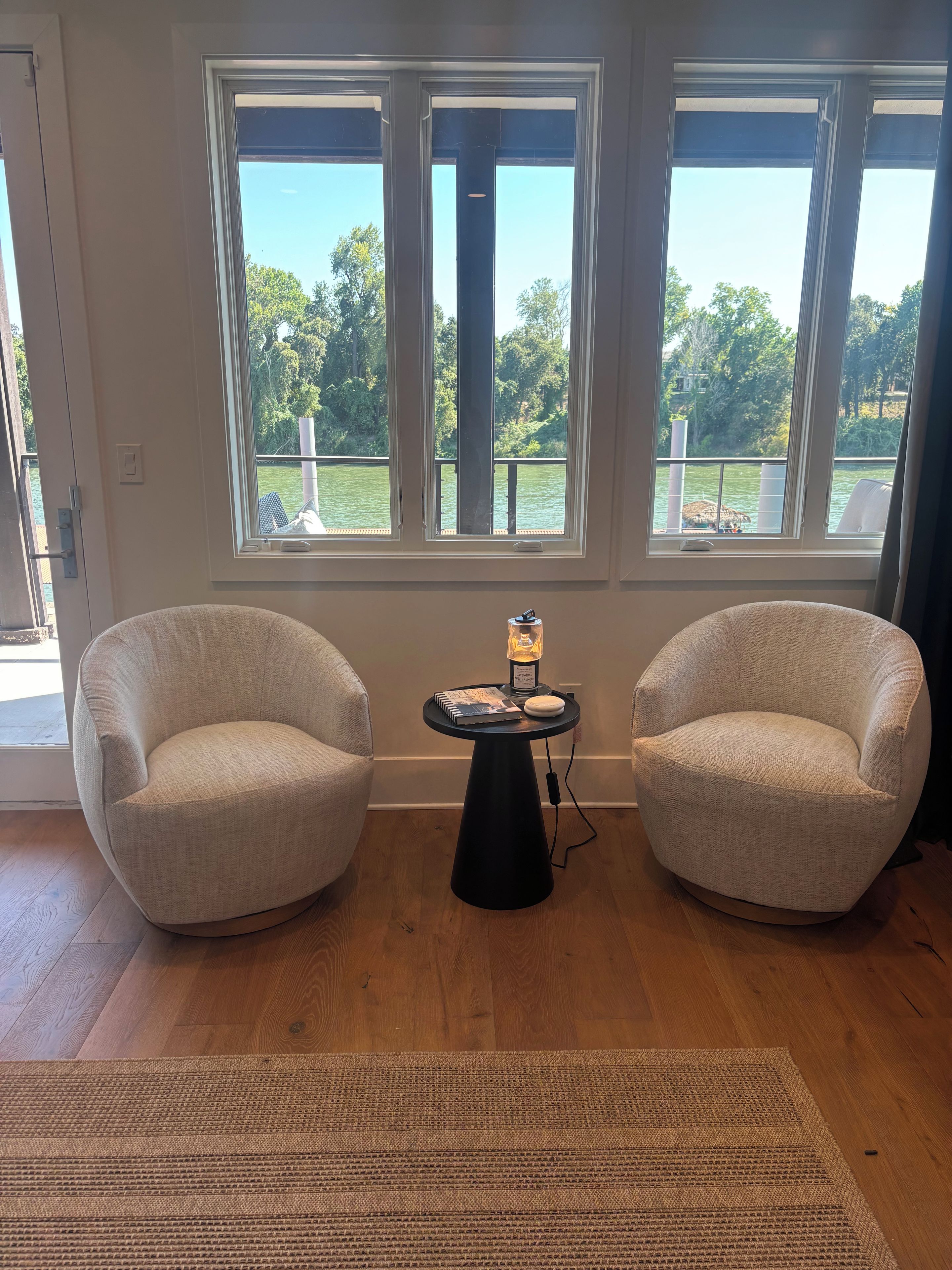 Une pièce comprenant deux fauteuils, une petite table avec une lampe et de grandes fenêtres donnant sur une étendue d’eau.