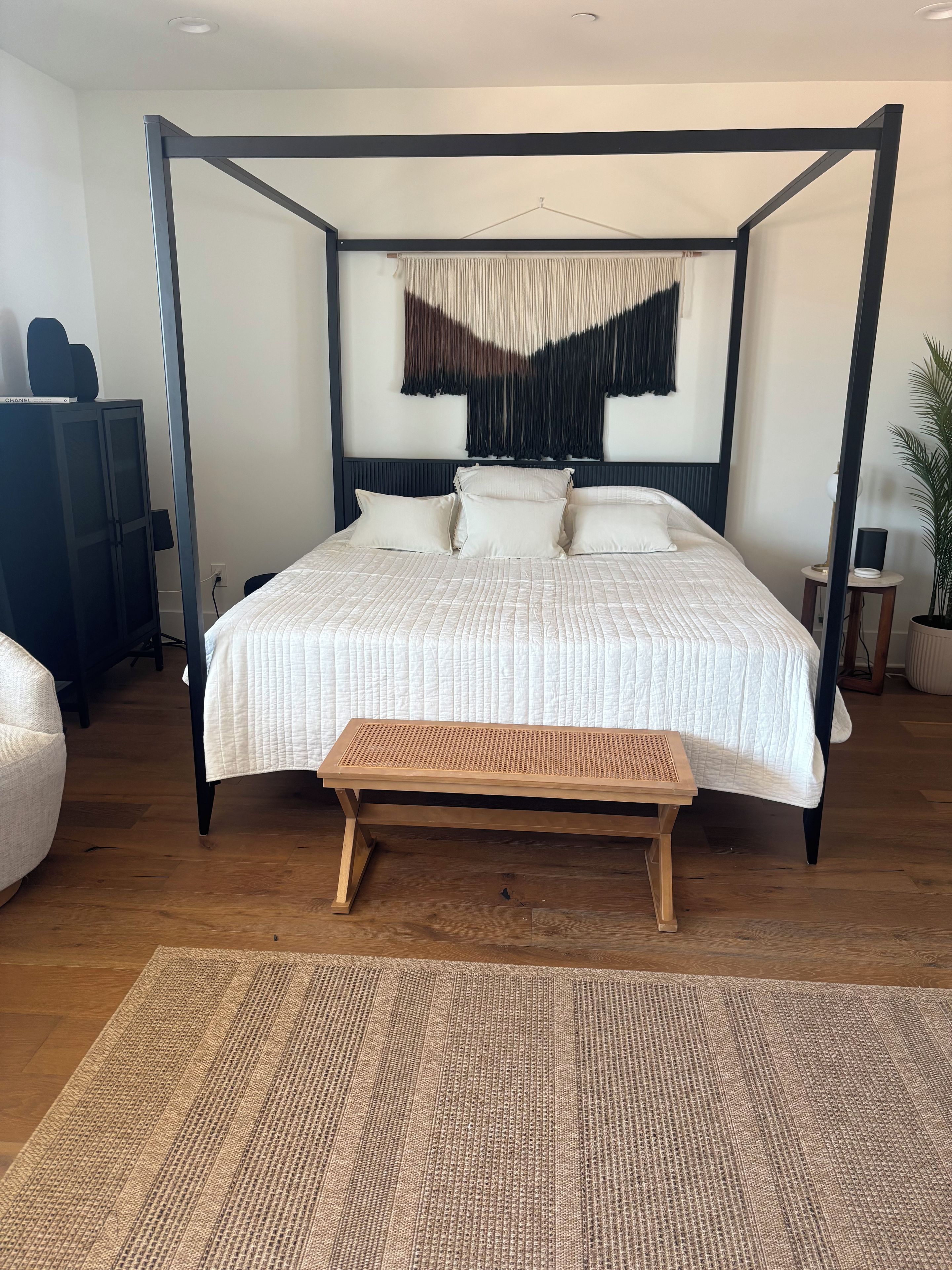 Un lit à baldaquin avec un banc en osier placé devant, un tapis en fibres naturelles et un haut-parleur fixé au mur.