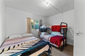 Ein Zimmer mit zwei Betten, eines mit einem gemusterten Überwurf und das andere mit einer roten Decke. Es gibt ein Fenster mit Vorhängen, eine Tür und eine Leuchte an der Decke.