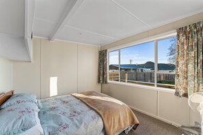 A bedroom with a large window, a bed with floral bedding, and a view of a beach and mountains. - The Little Jetty - Motuoapa Holiday Home (Motuoapa)