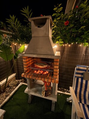 An outdoor barbecue grill with a chimney, surrounded by greenery and a decorative fence. - Garden & BBQ · Dinners Under the Porch Lights (Gran Alacant)