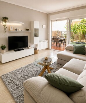 A modern living room with a flat-screen TV, a glass coffee table, a sofa, and a dining area visible through a glass door. - Garden & BBQ · Dinners Under the Porch Lights (Gran Alacant)