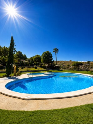 A clear blue swimming pool with a sunlit sky, surrounded by well-maintained grass and trees. - Garden & BBQ · Dinners Under the Porch Lights (Gran Alacant)
