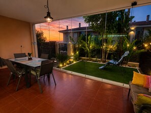 Une terrasse avec une table et des chaises, une petite pelouse et une vue sur une maison à travers un mur de verre.