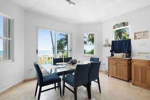 A dining area with a glass table and chairs, a kitchen with wooden cabinets, and a view of the ocean through large windows. - Cielo y Mar Beach House | Direct Beach Access (Humacao)