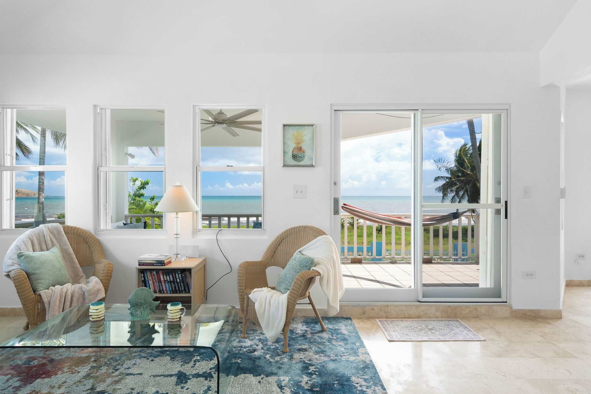 A living room with a glass table, two wicker chairs, a bookshelf, and a view of the ocean through large windows.