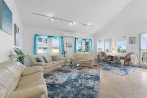A spacious living room with two sofas, a coffee table, and a chair. There are large windows with curtains, a view of the outdoors, and a door leading to a balcony. - Cielo y Mar Beach House | Direct Beach Access (Humacao)
