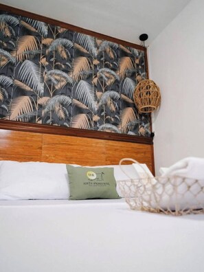 A hotel room with a wooden headboard, a tropical leaf-patterned wallpaper, a woven basket, and a pillow with a logo.